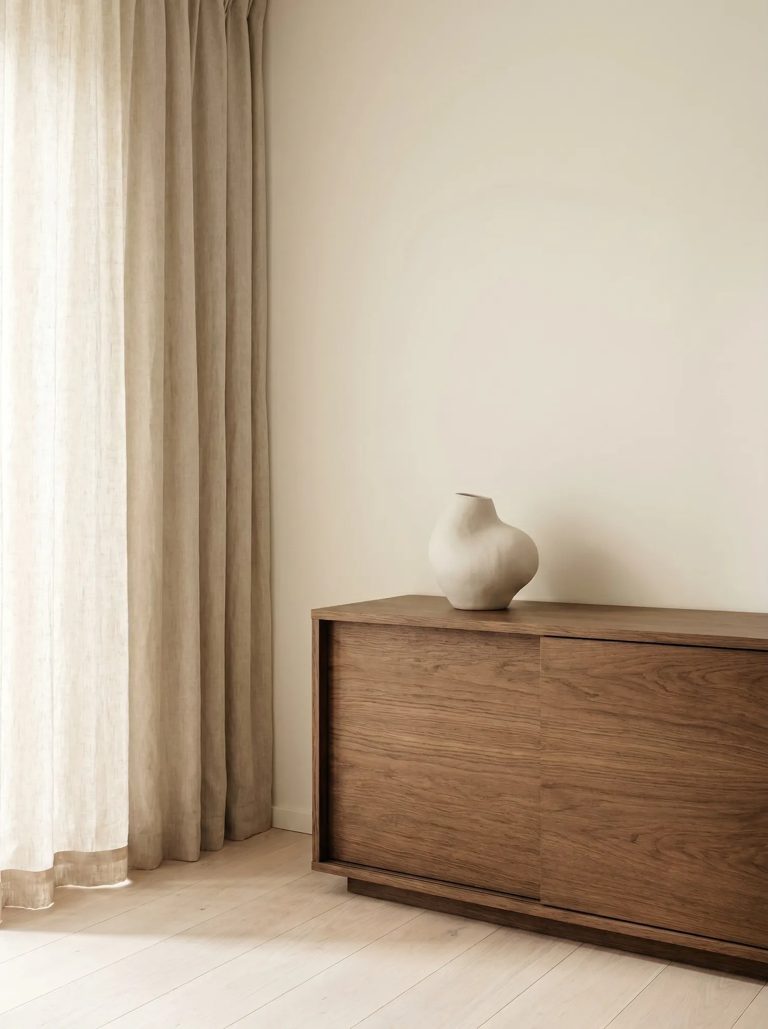 Calm interior corner with oak sideboard, ceramic object, linen curtain, and soft neutral walls